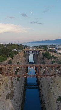 Vertical drone shot above Corinth Canal at sunset, Greece. Bridges visible, cars crossing. Clear day, few clouds. Golden light on narrow waterway