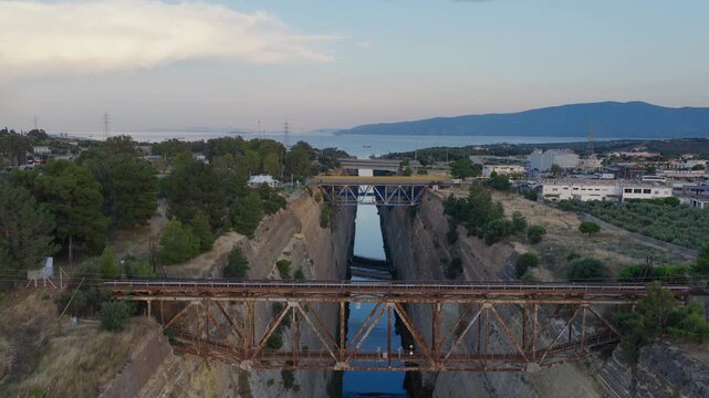 Drone shot above Corinth Canal at sunset, Greece. Bridges visible, cars crossing. Clear day, few clouds. Golden evening light on narrow waterway