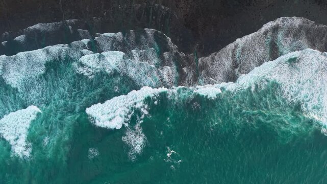 Aerial view of powerful ocean waves crashing against the rugged limestone cliffs and dark rocky shoreline of Gunungkidul Regency, Yogyakarta, Indonesia.