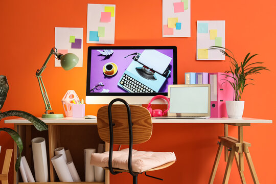Creative workspace and remote work environment, modern home office with computer and laptop on desk against orange wall for freelance professional