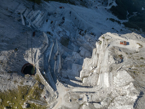 Aerial view of the Punta Carina marble quarry featuring terraced excavation, white marble rock faces, winding dirt roads, heavy machinery, and a small dark pool in Minucciano, Toscana, Italy.