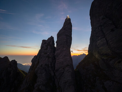 Aerial view of a climber rappelling down the sharp rock pinnacle of Punta Carina at twilight with a distant town glowing in the valley in Minucciano, Toscana, Italy.