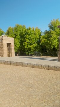 A wide shot of the Temple of Debod, an ancient Egyptian monument located in Madrid, Spain, surrounded by lush green trees under a clear blue sky.