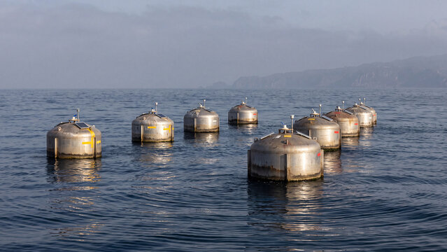 Steel wave energy buoys with solar sensors floating on calm sea near misty coastal cliffs. Offshore renewable ocean power farm harvesting wave motion for clean sustainable marine electricity