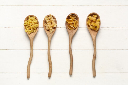 Wooden spoons with different types of pasta on white wooden background