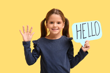Cute little happy girl holding paper speech bubble with word HELLO while waving hand on yellow background © Pixel-Shot