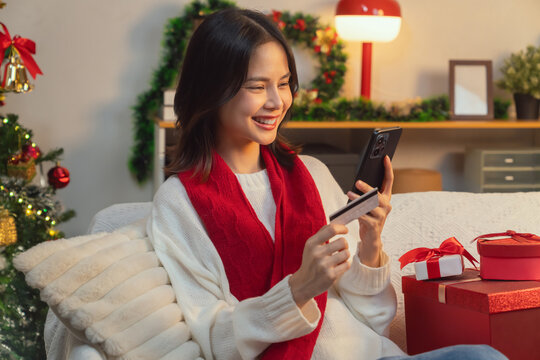 Cute Asian young woman wearing a white knitted sweater and red scarf, sitting on a sofa in the living room while using a smartphone and credit card for online shopping in Christmas day.