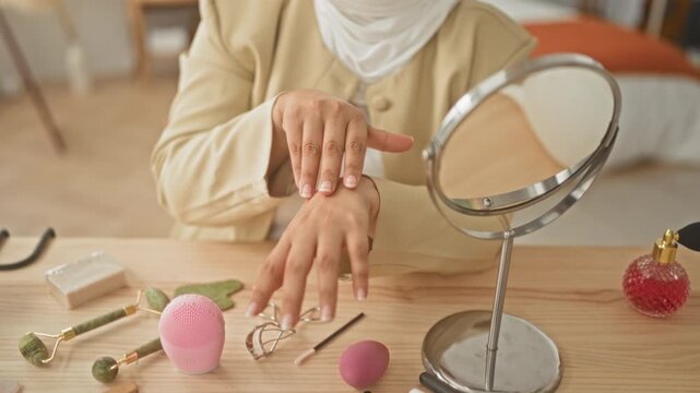 Woman wearing headscarf rubbing hands over moisturiser and skincare tools beside a tabletop mirror in studio; calm self care ritual.