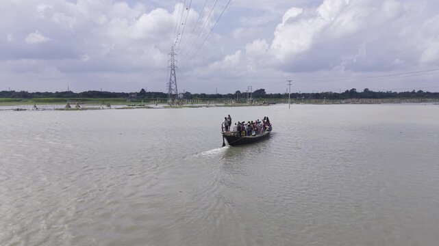 Aerial drone footage captures a passenger boat carrying people across the Padma River in Bangladesh, with riverside vegetation, an embankment, transmission towers, blue sky and scattered clouds.