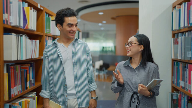 A young man and a woman with glasses walk through a library aisle together, chatting happily while holding books