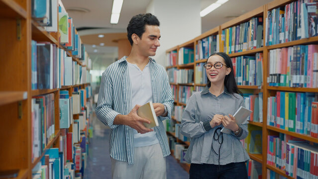 A young man and a smiling woman wearing glasses walk along a library aisle, chatting warmly while holding books between bookshelves