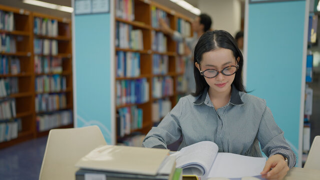 A young woman with glasses sits at a library table, quietly reading an open book with bookshelves filled with books behind her