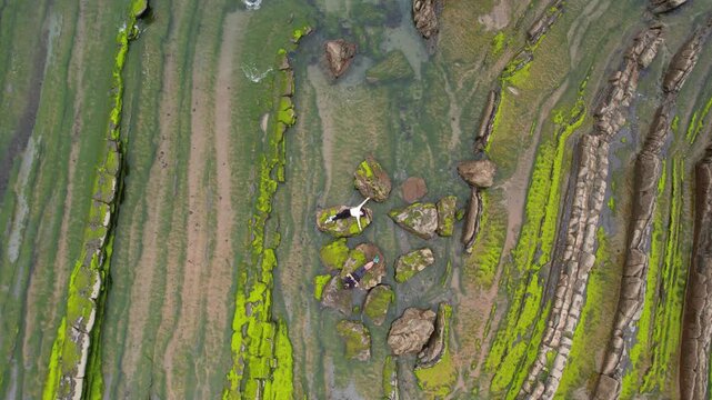 Aerial view of two people resting on mossy rocks along the coastal flysch formations in Bilbao, Basque Country, Spain.