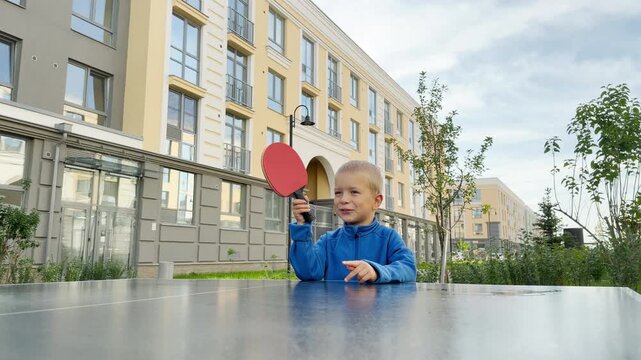 A happy young boy raises a red table tennis paddle with excitement. He plays ping pong in an outdoor courtyard near modern buildings. His bright smile brings pure joy and enthusiasm.