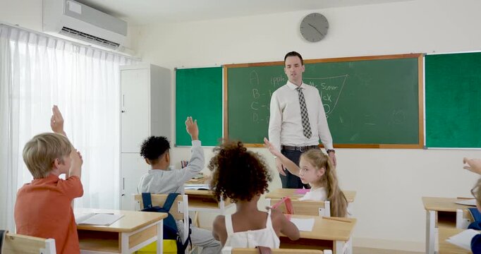 Male teacher asks math question near chalkboard while elementary pupil group eagerly raises arms, back to school discussion builds participation, confidence, classroom discipline, learning success