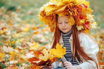 Close up of a redhead girl in a leaf crown sitting on the ground looking down at a yellow maple bouquet. © Dmytro