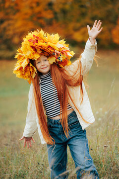 Smiling redhead girl in a leaf crown waving her hand on a grassy hill with blurred warm autumn forest background.