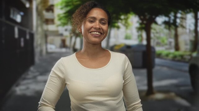 Woman smiling and giving a thumbs up with raised hand on a city street near trees and buildings; happiness.