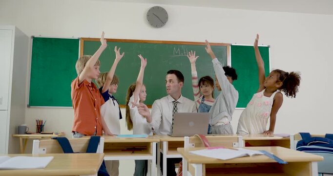 Male adult teacher gives highfive while young multicultural student group raises hand near chalkboard, celebrating answer confidence during first day elementary classroom discussion and learning tasks