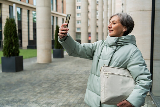 Woman with grey hair taking a selfie with her smartphone outdoors. Senior lady holding a bag in city