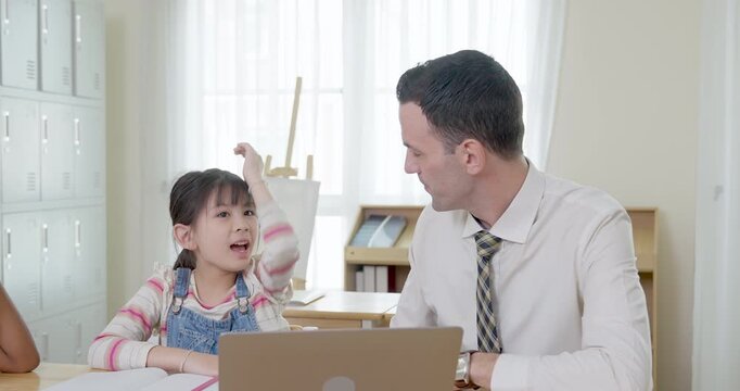 Adult male teacher listens as elementary girl raises arm and answers question beside laptop, back to school tutoring scene builds confidence, communication, participation, readiness, trust, respect