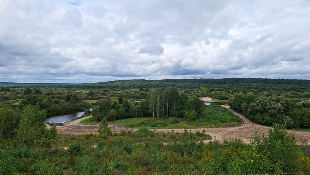 Expansive natural landscape with winding dirt roads and serene river under cloudy skies
