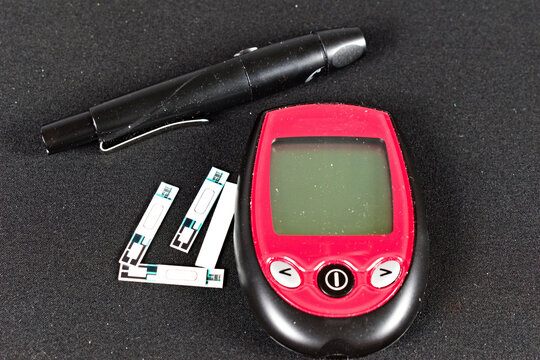 A red and black glucose meter with a test strip inserted sits next to a black lancet pen on a textured dark surface.