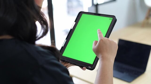 Back view of Asian Woman sit use digital tech tablet computer open green screen mock up at home