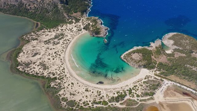 Drone shot captures the iconic Omega-shaped Voidokilia Beach in Pilos, Messinia, Greece. Crystal blue sea, white sands, coastal dunes, vivid lagoon, and natural scenery in bright daylight.