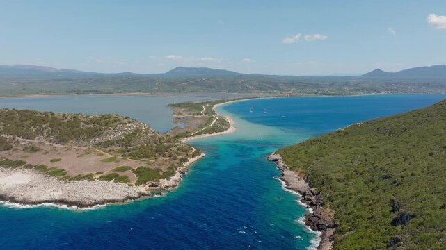 Drone video captures crystal clear turquoise waters, unique sandbar, vibrant coastal scenery and lush greenery at Sykia and Chrisi Ammos beaches, Pilos, Greece on a sunny day.