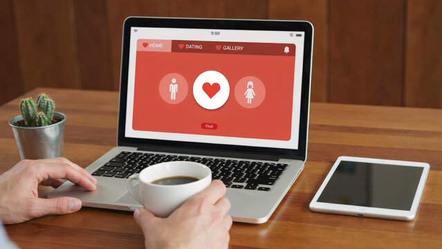 Laptop displaying dating app interface with heart icon and user profiles on a wooden desk with coffee