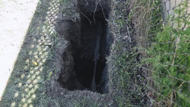 A directional drill bit pulls streetlight conduit back through an underground bore as the trench exposes moist soil, buried pipe, and the active pathway beside a fence line.