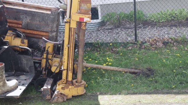 A hydrovac machine uses a drill rod to bore into the ground beside a fence, creating a subsurface path for a streetlight pole installation; hydraulic arm, drill rod, and grassy work area are visible.