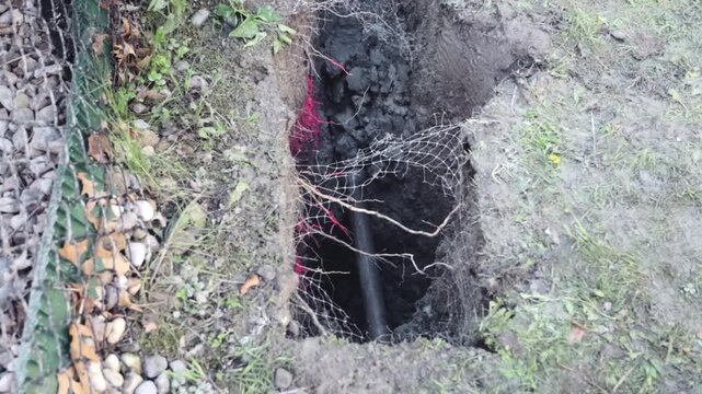 A directional drill bit pushes through a test pit as dark soil, trench walls, buried pipe, mesh wrap, and marked ground frame the active pathway for underground streetlight installation.