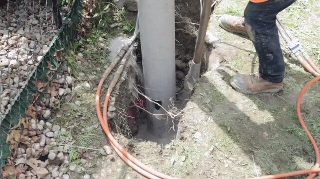 A crew member uses a hydraulic compactor to press soil around the base of a newly installed streetlight pole as orange cables, exposed earth, mesh wrap, and a digging tool frame the active foundation