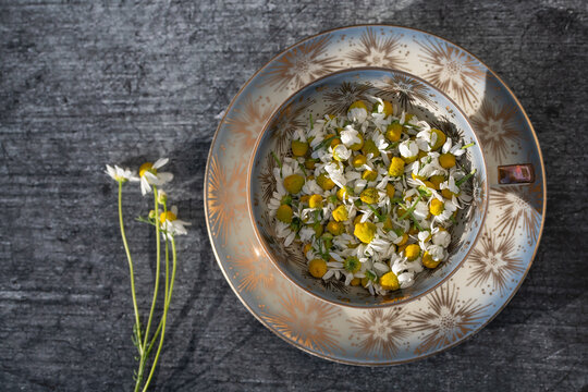 A close-up of an old teacup filled with chamomile flowers. A dark table is visible in the background. The cup is photographed from above.