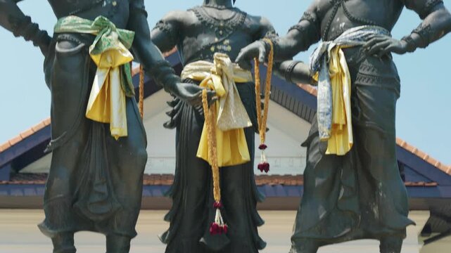 Low angle shot of the three bronze statues of Lanna kings at the Three Kings Monument with the Thai national flag fluttering in the background against a clear sky in Chiang Mai, Thailand.