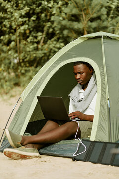 Young African American man sitting in camping tent outdoors, working on laptop powered by portable solar panel. Image suitable for digital nomad lifestyle, vertical