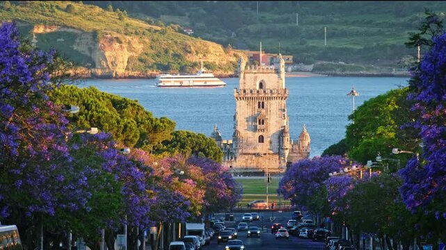 Jacaranda trees bloom with purple flowers near Belem Tower during sunset in Belem, Lisbon, Portugal. The scene shows the beauty of spring by the river