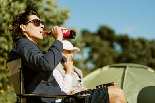 Young adult Caucasian man sitting in folding chair drinking beverage during camping trip outdoors. Visual suitable for outdoor lifestyle branding, travel campaigns and summer recreation marketing