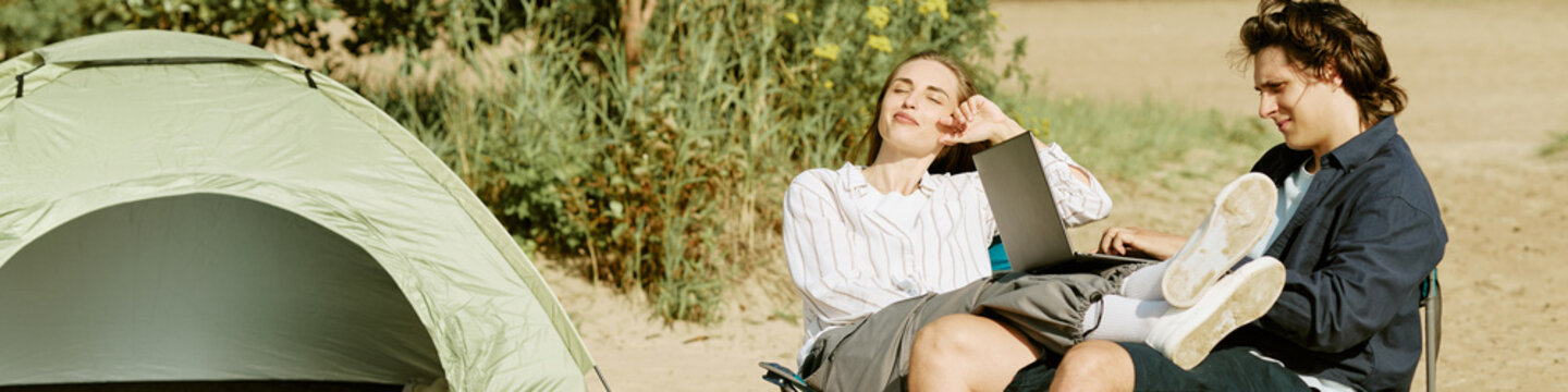 Header of young adult Caucasian couple relaxing by camping tent outdoors using laptop computer. Image useful for remote work freedom, digital nomad lifestyle and travel campaign advertising