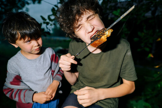 Teenager bites burnt toast outdoors while younger boy watches him