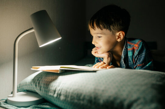 Smiling boy reads a book in bed at night
