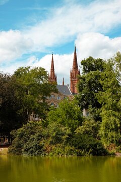 Wiesbaden, Hesse, Germany &ndash; Marktkirche church towers rising above lush greenery and pond in scenic urban park landscape.