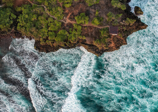 Aerial view of the rugged coastline of Gunungkidul Regency with crashing turquoise waves, rocky cliffs, and lush vegetation, Gunungkidul Regency, Special Region of Yogyakarta, Indonesia.