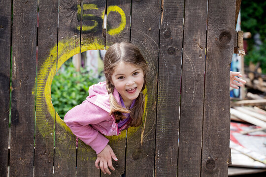 A happy girl peeks through a wooden hole at an adventure playground