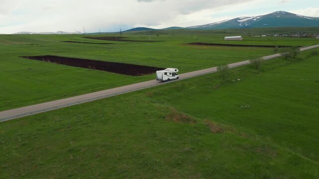 A white camper rides on an empty asphalt road through a vast green landscape with snow-capped mountains in the background, embodying the concept of freedom, travel and adventure vacation in Georgia
