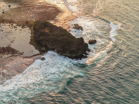 Aerial view of a rocky outcrop surrounded by crashing waves and turquoise water in Gunungkidul Regency, Special Region of Yogyakarta, Indonesia.