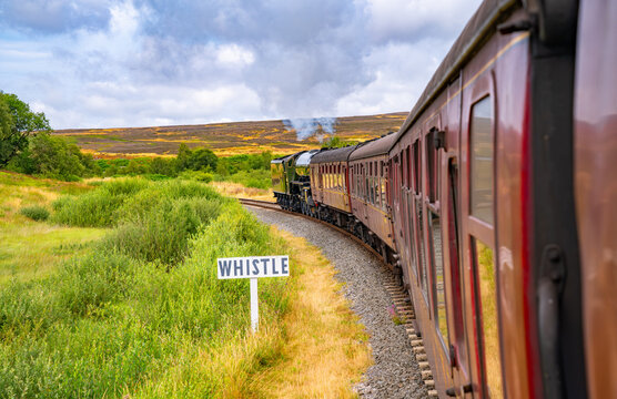 View along a historic steam train travelling through the open moorland of North Yorkshire, England, with a whistle sign beside the curved railway track.