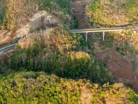 Aerial view of a concrete bridge spanning a lush forested valley with terraced fields and rocky cliffs in Gunungkidul Regency, Special Region of Yogyakarta, Indonesia.